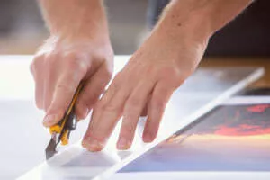 Weiterverarbeitung Fotodruck Print Moonfeld auf Leinwand, ein Mitarbeiter schneidet mit Cutter und Lineal den Großformatdruck auf Leinwand zurecht