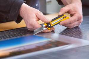 Weiterverarbeitung Fotodruck Print Landschaft Alu-Dibond Messer, ein Mitarbeiter bringt die Schutzfolie auf den Print auf und schneidet die Folie mit Cutter zurecht