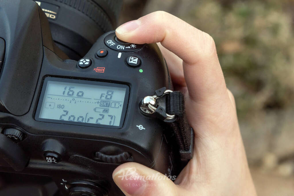 Eine Frau drückt den Auslöser einer Nikon Spiegelreflexkamera leicht. Das Display zeigt die Kamera-Einstellungen.