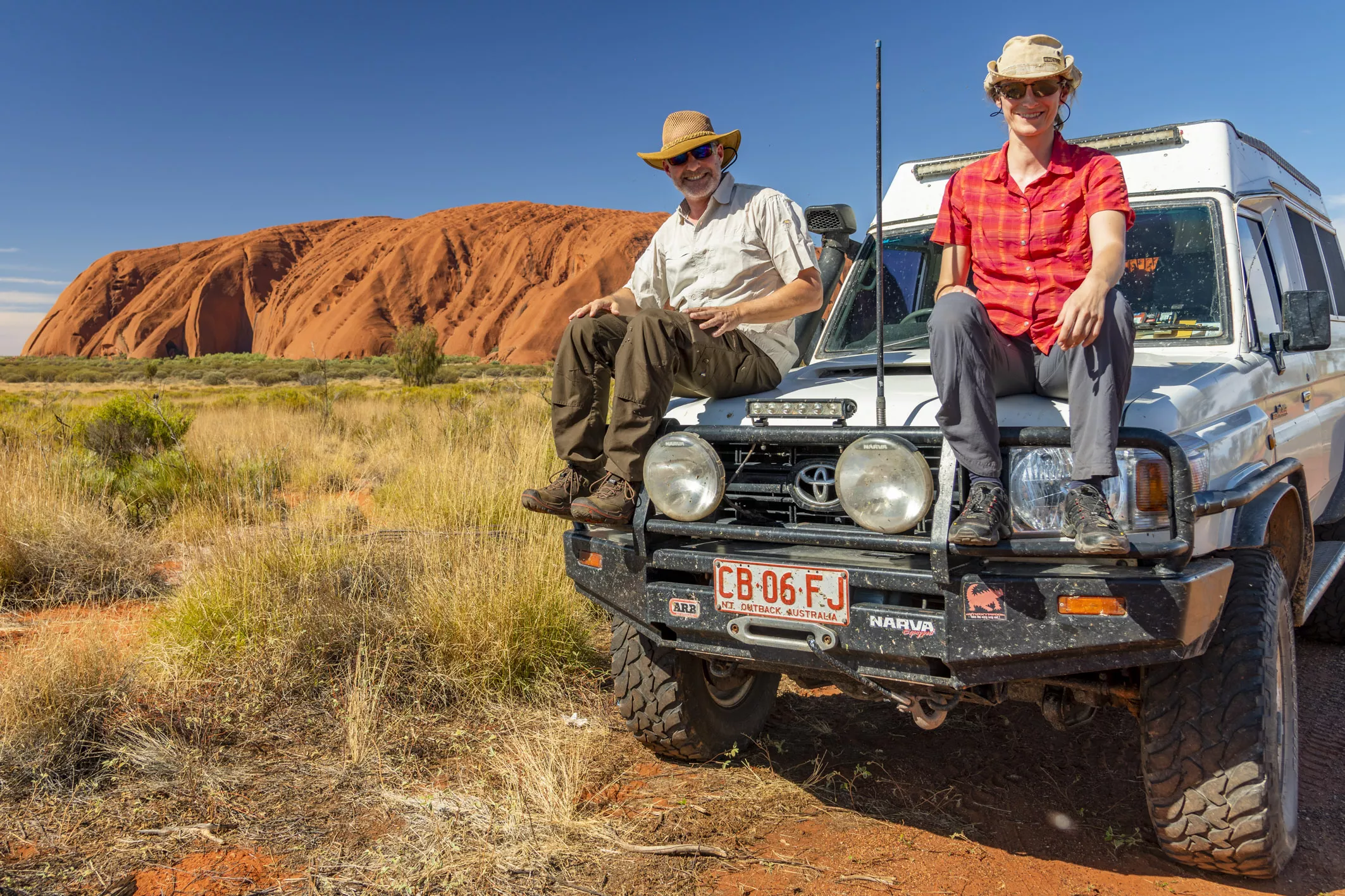 die Fotografen Aneta und Dirk Bleyer sitzen auf der Kühlerhaube eines Jeeps am Uluru in Australien