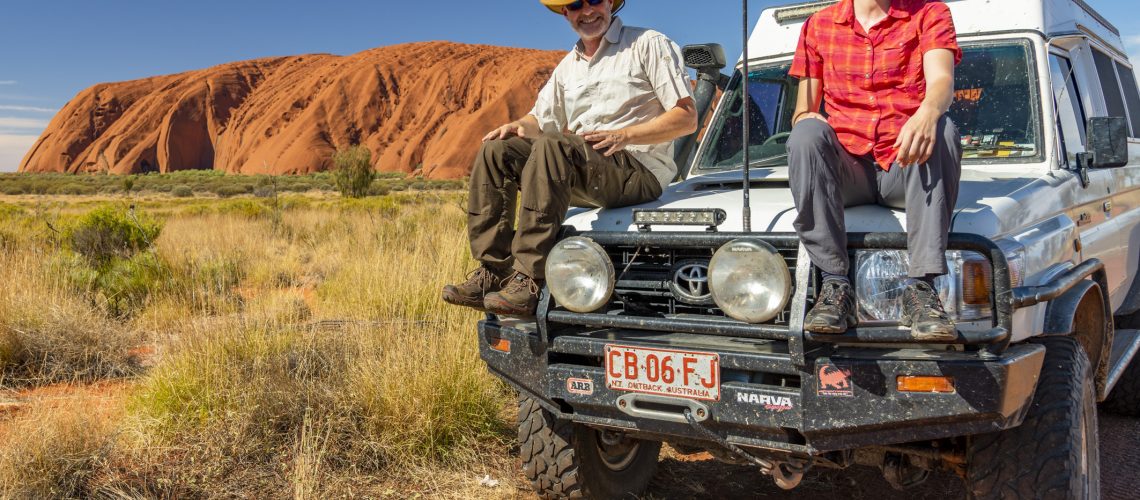 die Fotografen Aneta und Dirk Bleyer sitzen auf der Kühlerhaube eines Jeeps am Uluru in Australien