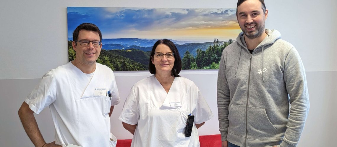 Oberarzt Dr. C. Häfele, Pflegeleitung Frau U. Stolzer und Geschäftsführer der Heimatlichter GmbH Benjamin Kuderer auf der Abteilung für Akutgeriatrie am Ortenau Klinikum in Offenburg vor einem Wandbild von Heimatfotos